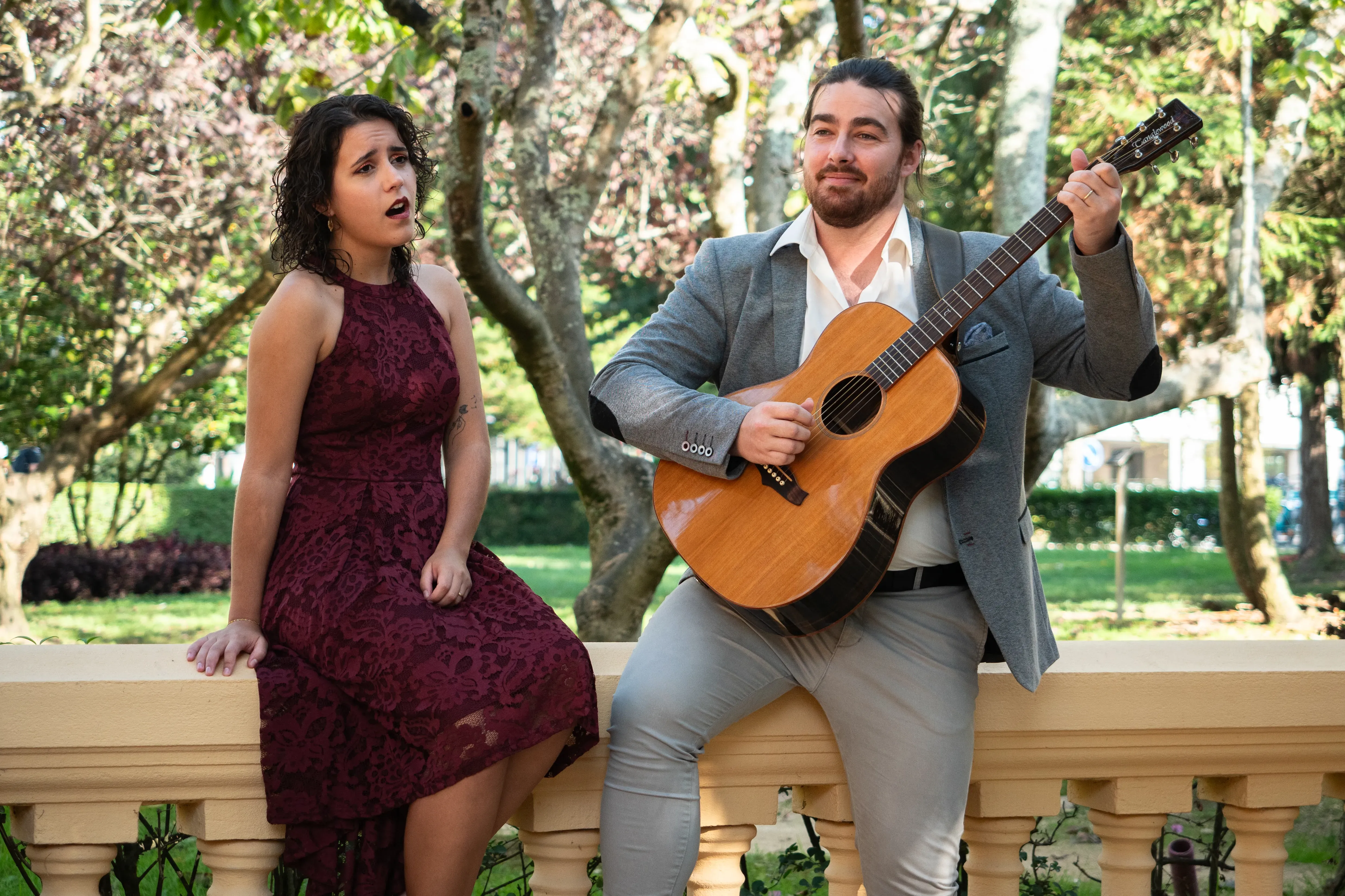 Um casal sentado no muro de um parque. A rapariga está a cantar e tem um vestido bordô e o rapaz tem um fato cinzento e está a tocar guitarra clássica.